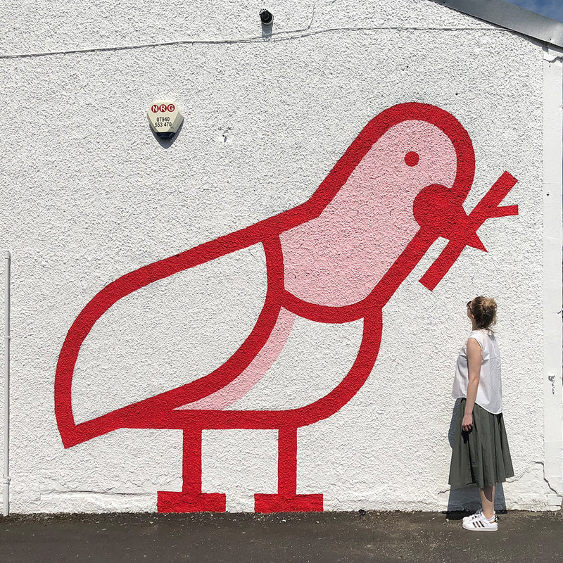 Large mural of a red pigeon on a white wall being looking at by a blonde woman